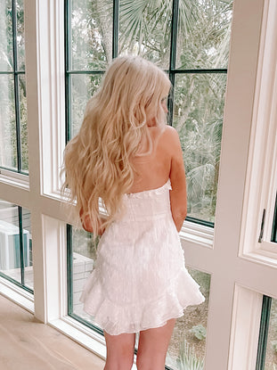 White eyelet mini dress with ruffled hem, shown from behind near a window.
