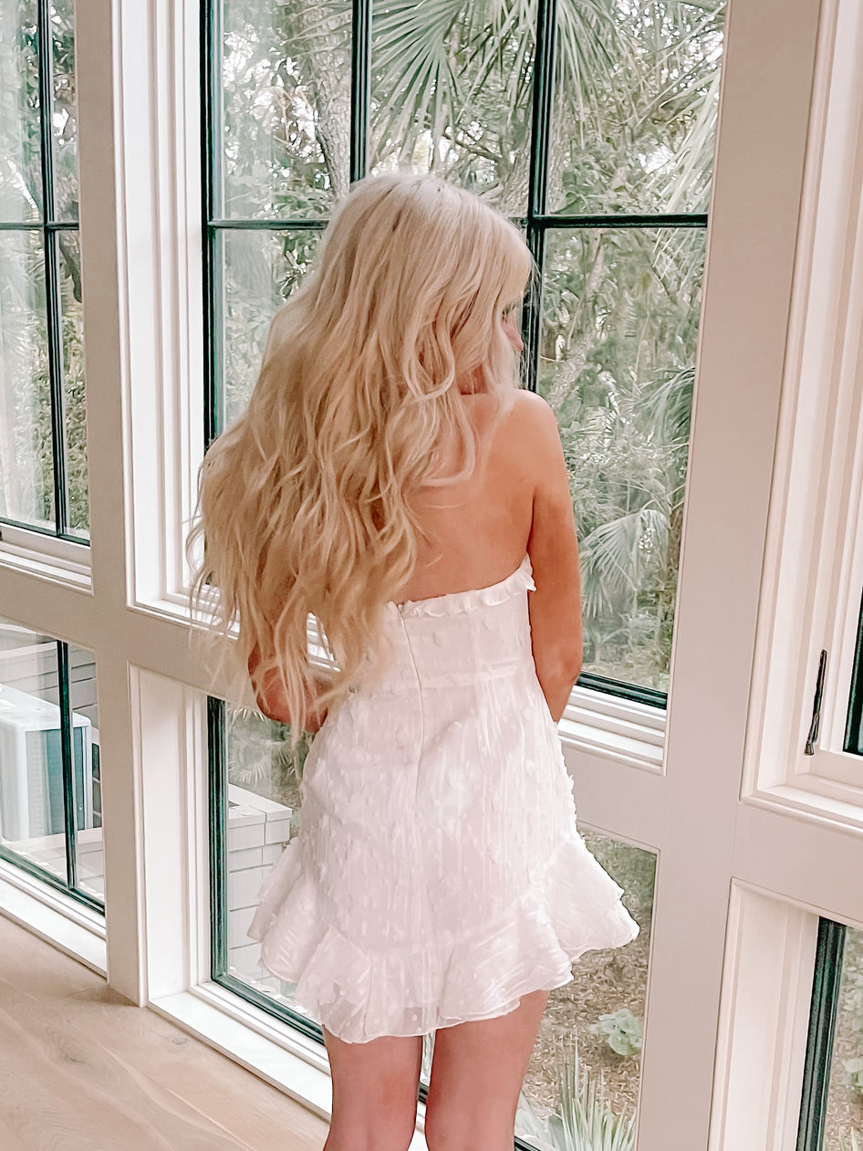 White eyelet mini dress with ruffled hem, shown from behind near a window.
