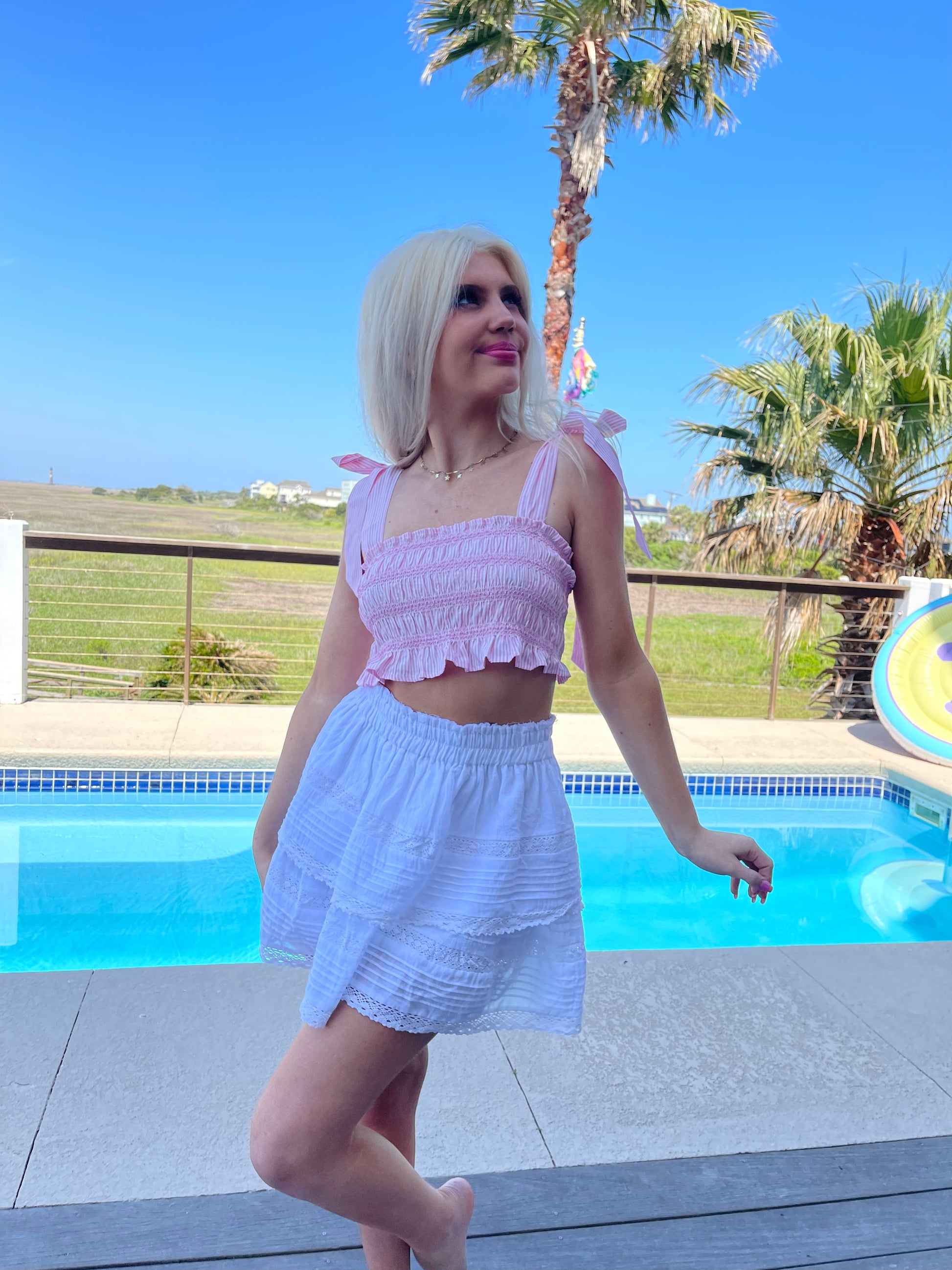 Woman by pool in pink crop top and white eyelet skirt.
