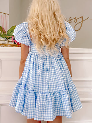 Blue gingham mini dress with puff sleeves and tiered skirt, shown from the back against a white wall.
