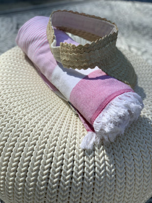 Straw visor and pink striped beach towel resting on a textured cream-colored surface.
