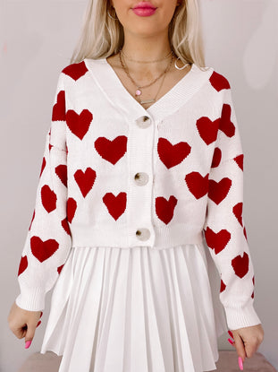 White cropped cardigan adorned with red hearts, button closure, modeled over a pleated white skirt.
