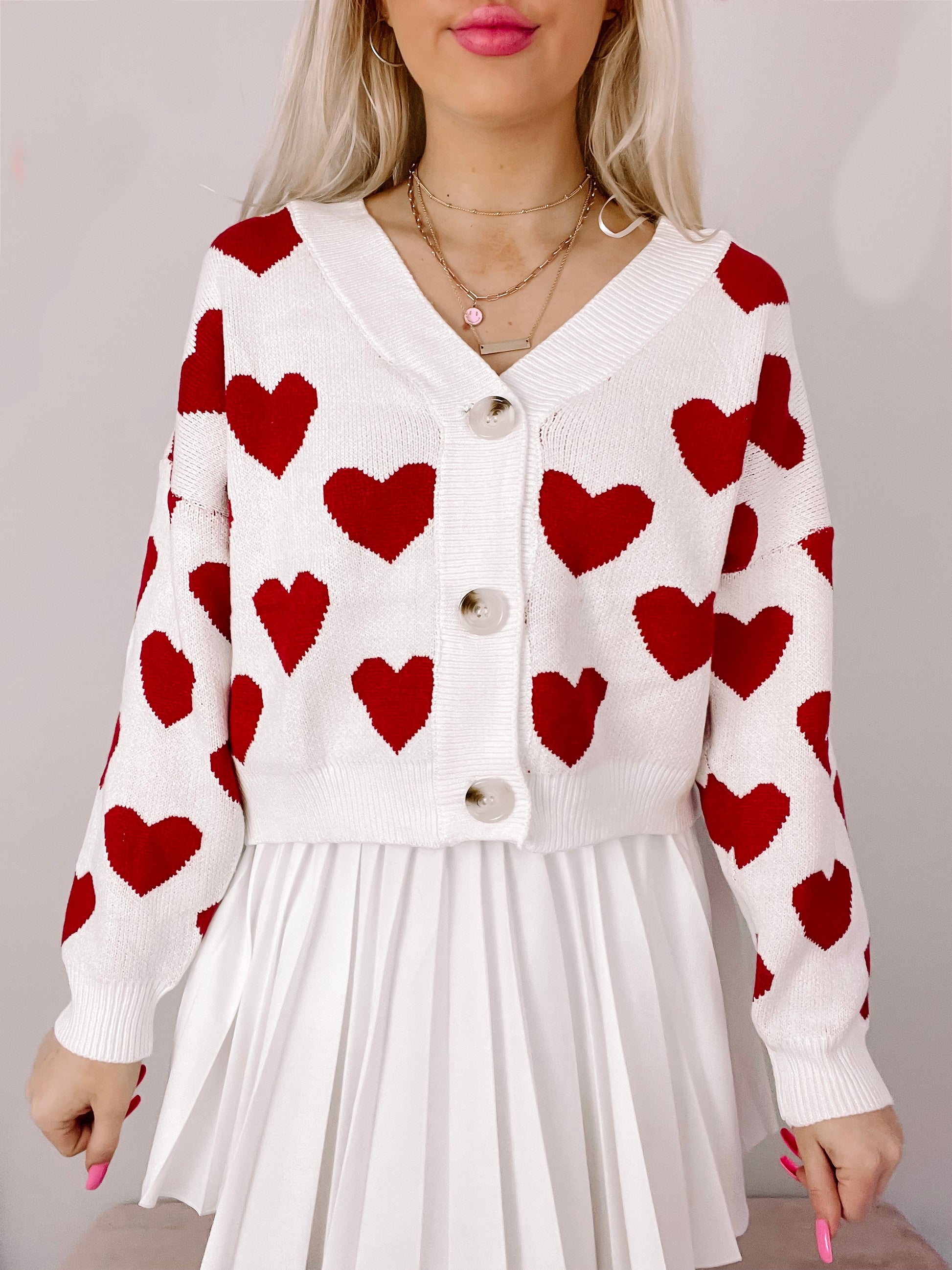 White knit cardigan adorned with red hearts, button closure, modeled over a pleated white skirt.
