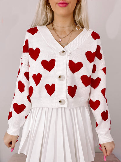 White knit cardigan adorned with red hearts, button closure, modeled over a pleated white skirt.
