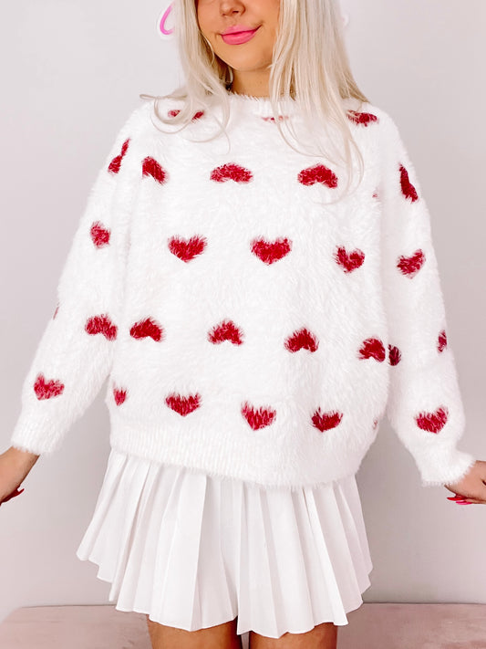Fuzzy white sweater adorned with small red hearts; styled with a white pleated skirt.