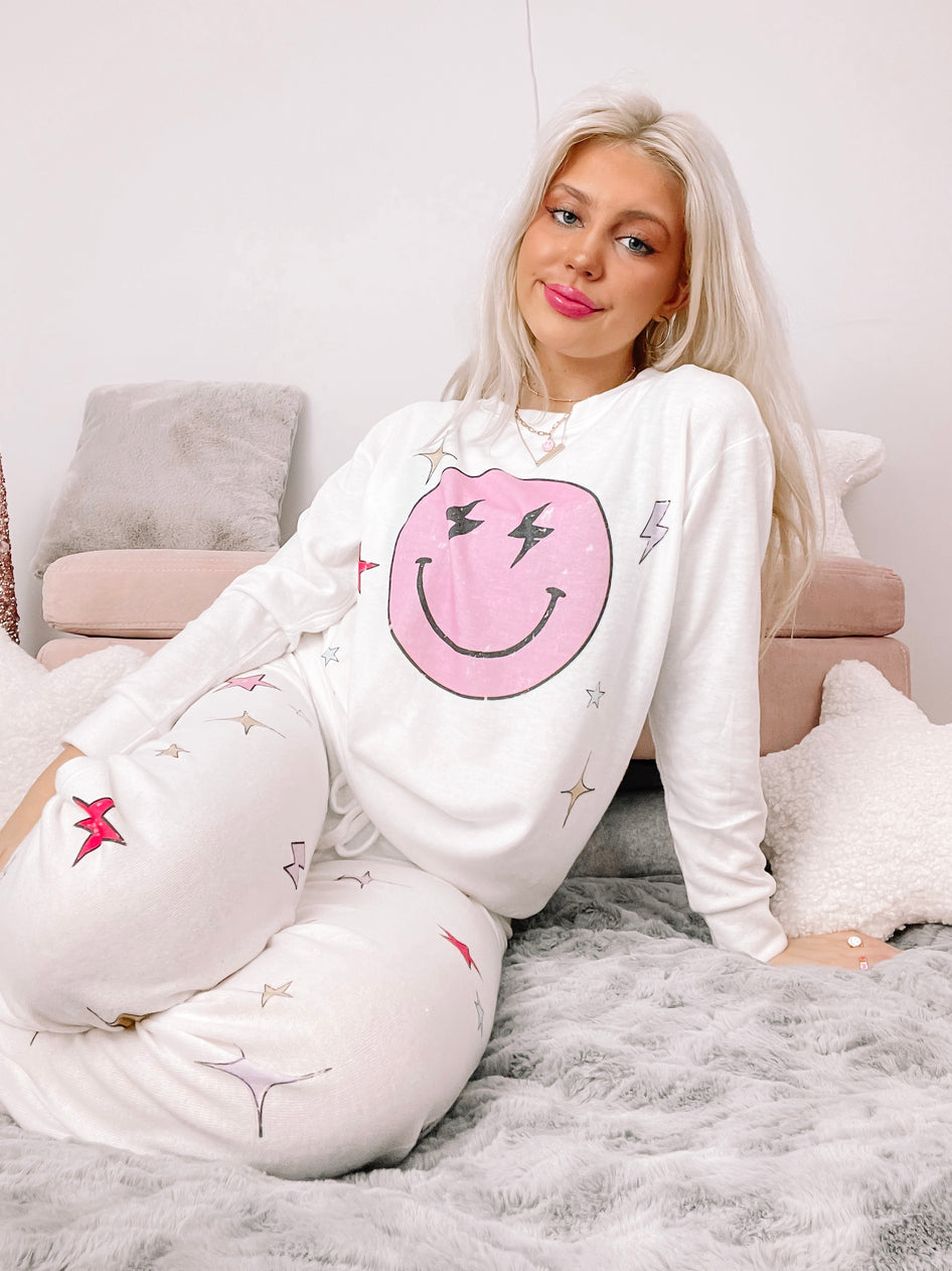 White pajama set with a pink smiley face and star print, modeled on a fluffy gray blanket.
