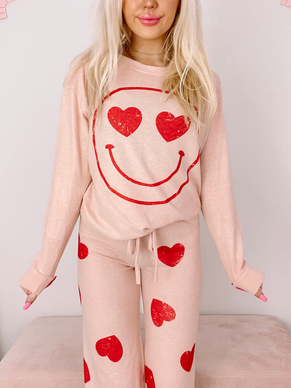 Smiley & Smitten PJ set: Blush pink lounge set with red heart print pants and a matching sweatshirt featuring a smiley face with heart eyes.
