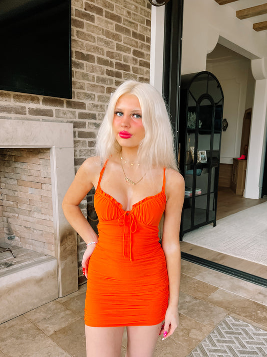 Blond woman in a vibrant orange mini dress, posing near a brick fireplace.