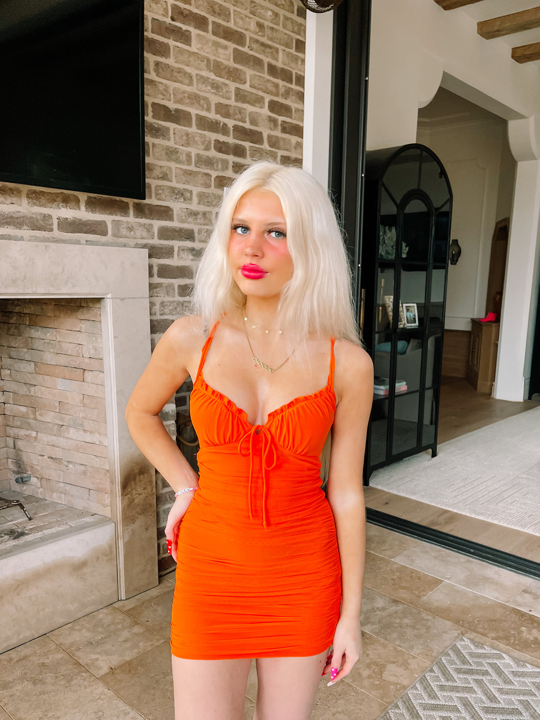 Blond woman in a vibrant orange mini dress, posing near a brick fireplace.

