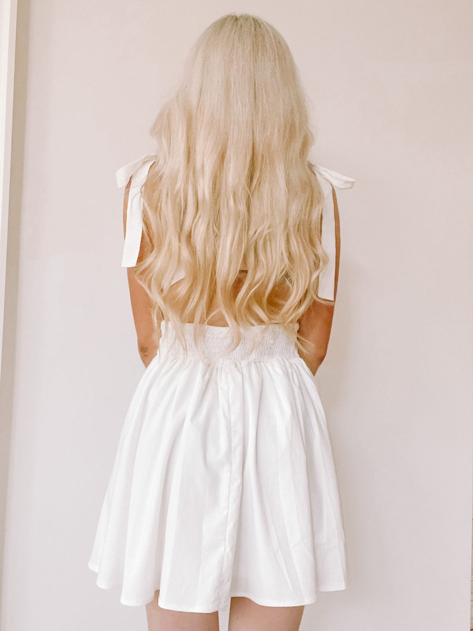White eyelet mini dress with bow shoulder straps, shown from the back against a neutral backdrop.
