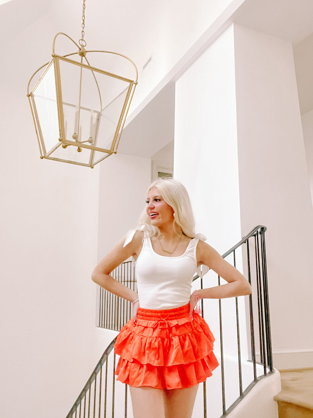 Bright orange tiered ruffle mini skirt styled on a model in a white tank top, near a staircase.

