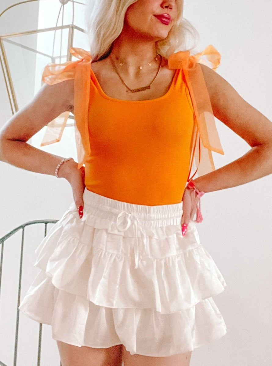 Orange bodysuit with sheer orange ribbon shoulder ties, styled with a white ruffled mini skirt.
