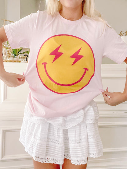 Pink t-shirt featuring a yellow smiley face with pink lightning bolts for eyes; shown on model.
