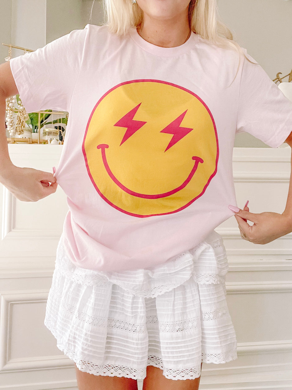 Pink t-shirt featuring a yellow smiley face with pink lightning bolts for eyes; shown on model.
