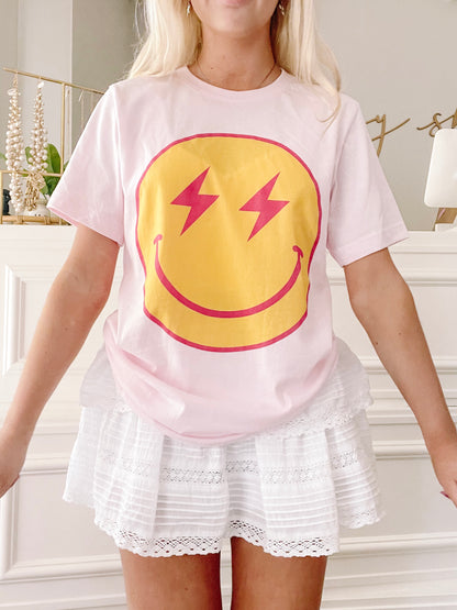 Pink t-shirt featuring a yellow smiley face with red lightning bolt eyes;  model wearing white eyelet shorts.
