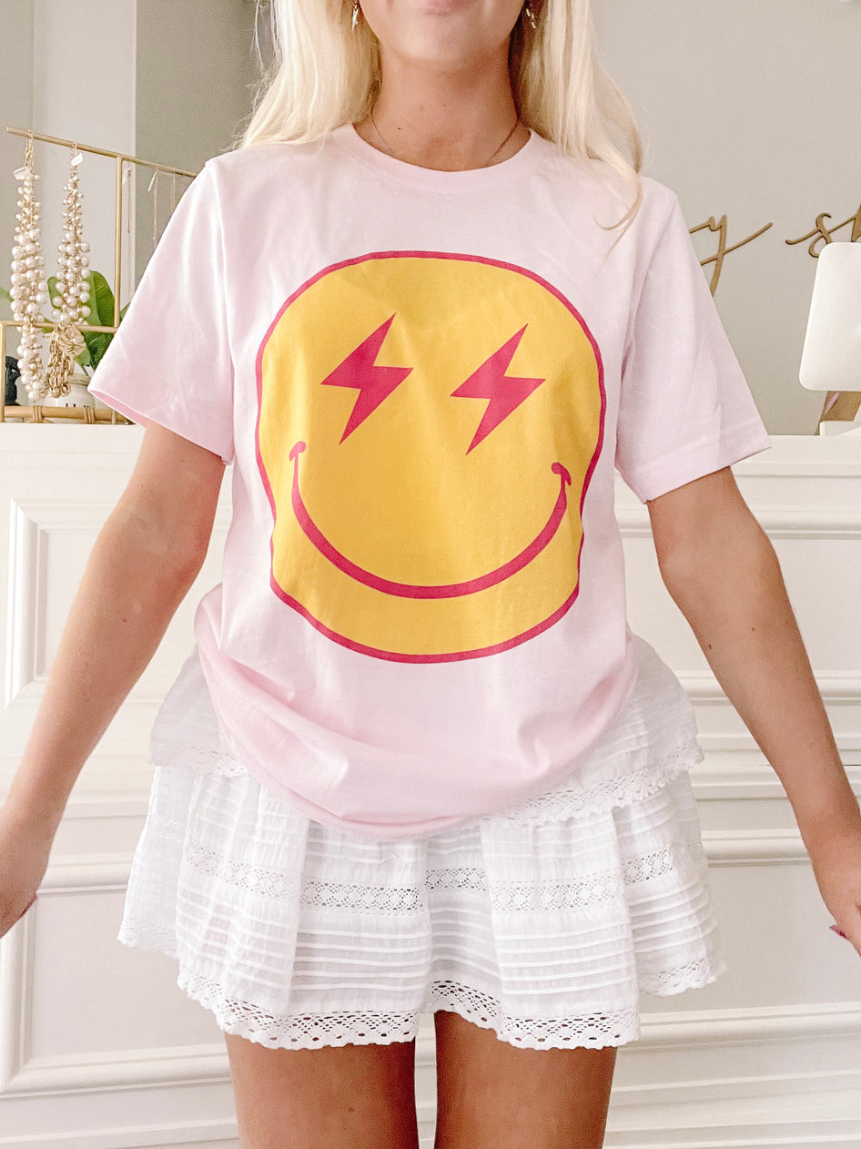 Pink t-shirt featuring a yellow smiley face with red lightning bolt eyes;  model wearing white eyelet shorts.
