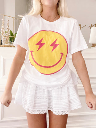 White cotton Striking Smiles tee featuring a yellow smiley face with pink lightning bolt eyes.
