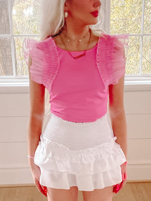 Hot pink ruffle top with sheer sleeves, styled with white ruffled shorts near a window.
