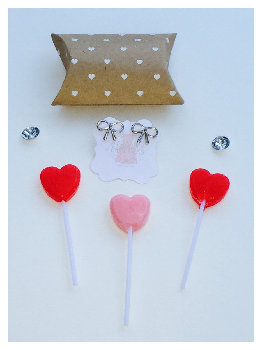 Silver bow earrings displayed with heart-shaped lollipops and kraft paper packaging.
