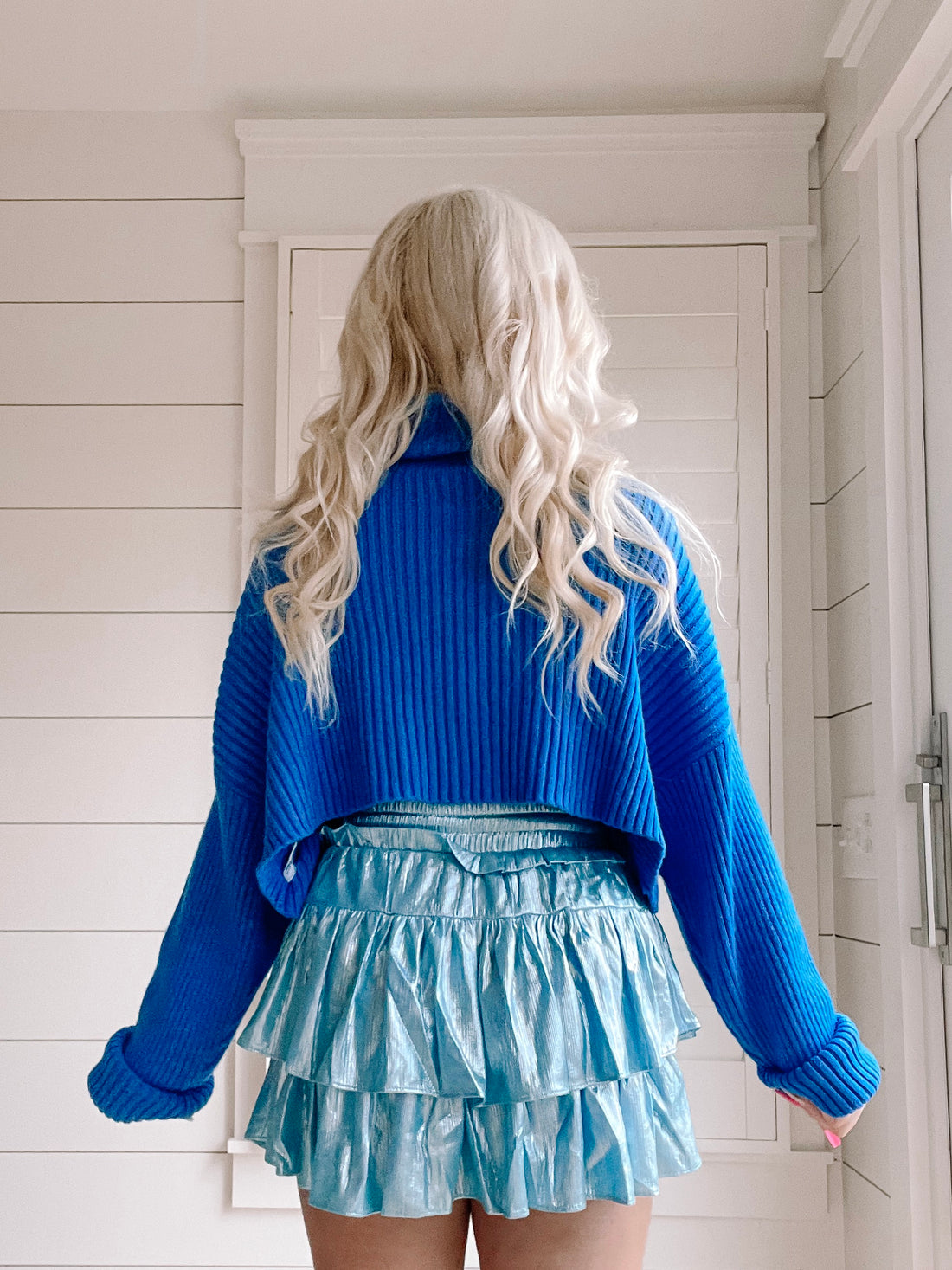 Bright blue cropped knit sweater with cuffed sleeves, shown from the back against a white shiplap wall.
