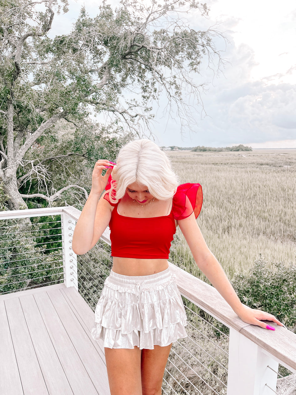 Red crop top with sheer flutter sleeves, styled outdoors on a deck overlooking a marsh.
