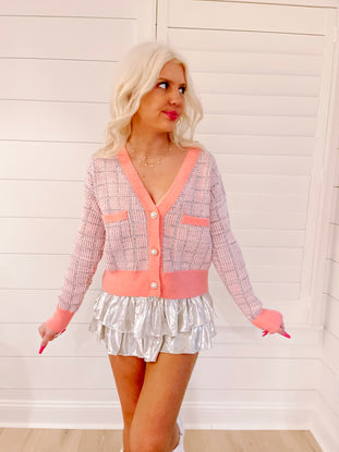 Pink and gray patterned cardigan with coral trim, button closure, and a model wearing it against a white paneled wall.
