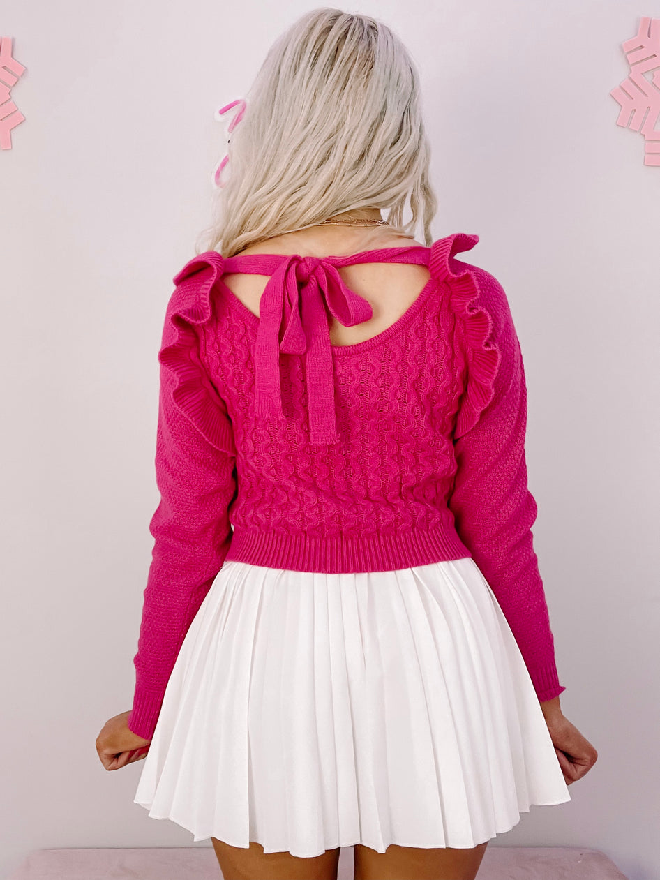 Hot pink cable-knit sweater with ruffle detailing and tie back, paired with a white pleated skirt.
