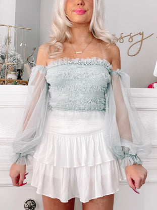 Sheer Luck Top: Light green off-the-shoulder smocked crop top with sheer long sleeves and ruffled trim, paired with a white tiered ruffle mini skirt.
