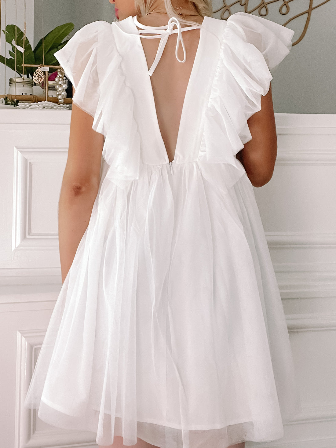 White eyelet mini dress with flutter sleeves and open back, shown from behind against a white paneled wall.
