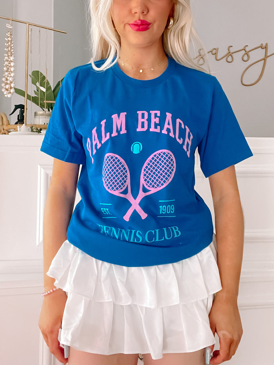 Bright blue tee featuring pink tennis racquets and "Palm Beach Tennis Club" graphic; worn with white ruffled skirt.
