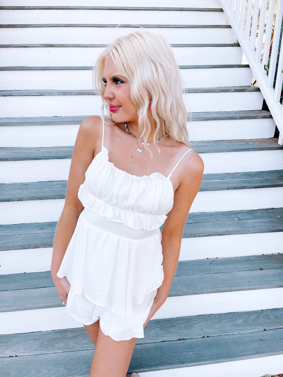 White ruffled romper on a model, posing on outdoor stairs.
