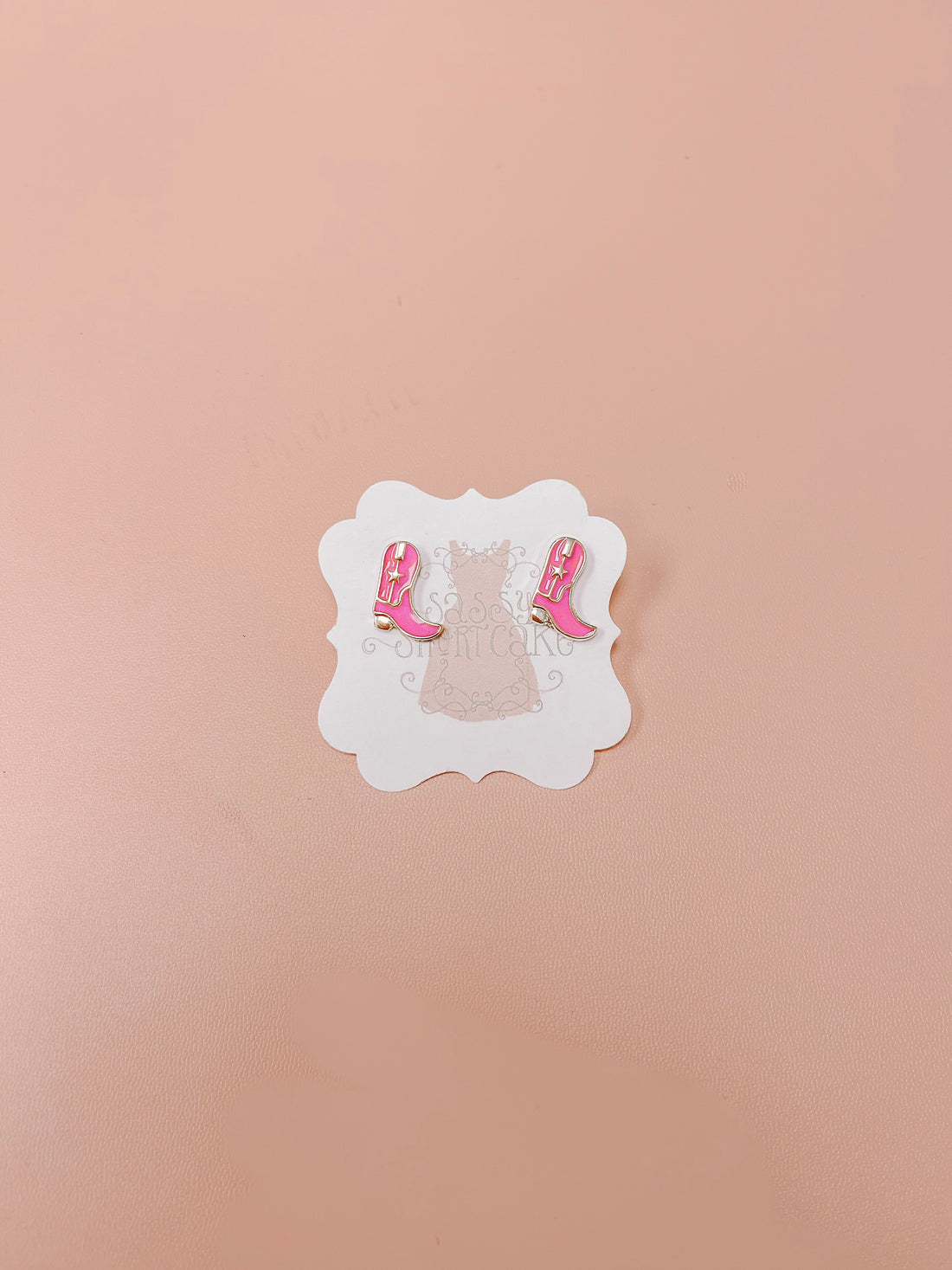 Pink cowboy boot stud earrings displayed on a light pink background.

