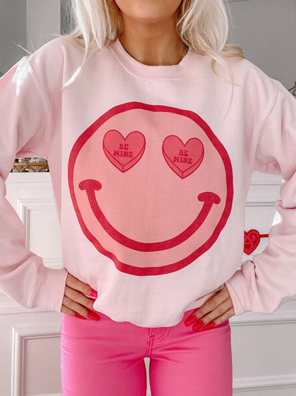 Pink crewneck sweatshirt featuring a smiling face design with "Be Mine" candy hearts for Valentine's Day.
