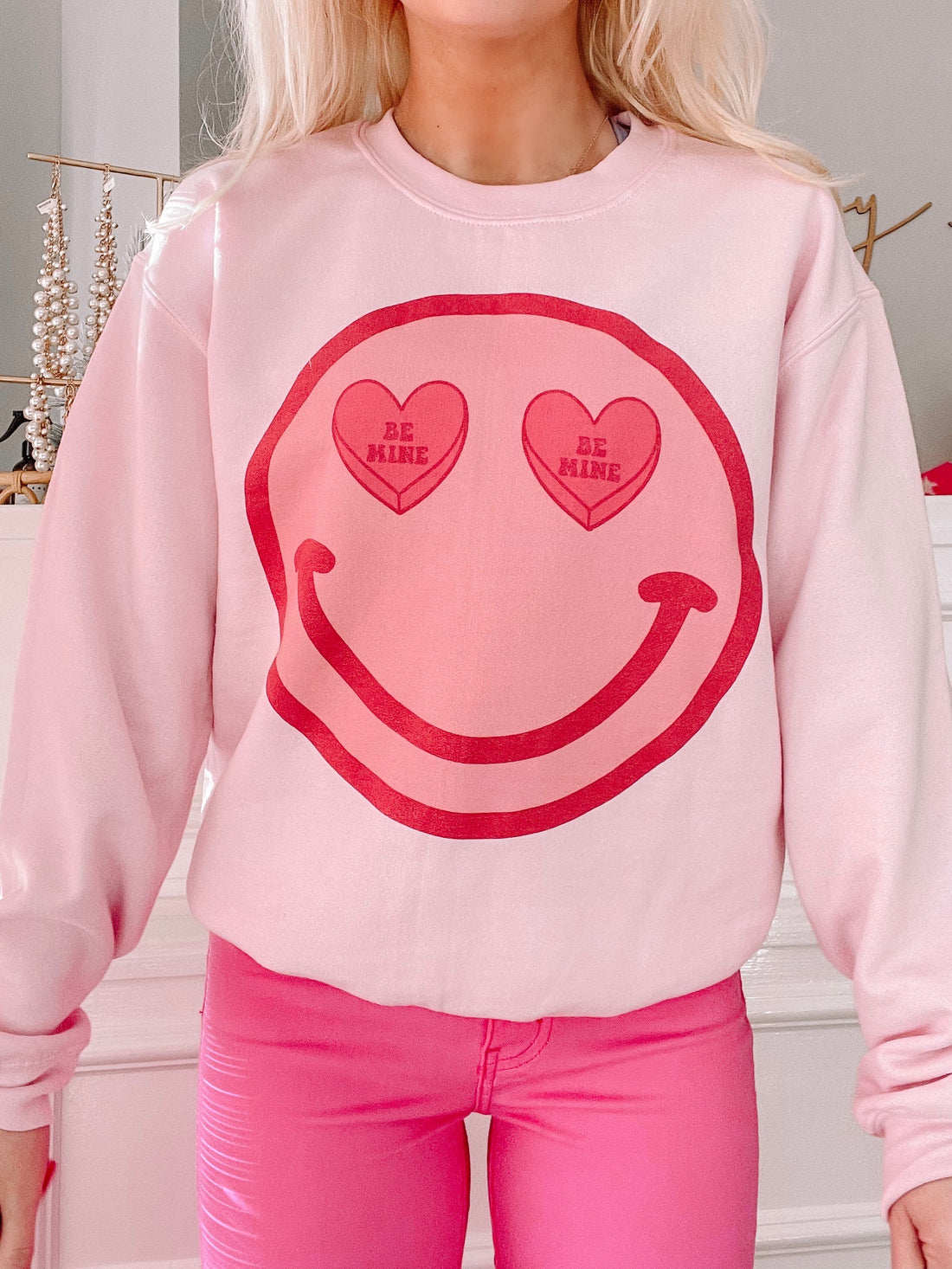 Pink crewneck sweatshirt featuring a smiling face graphic with "Be Mine" candy hearts for eyes.
