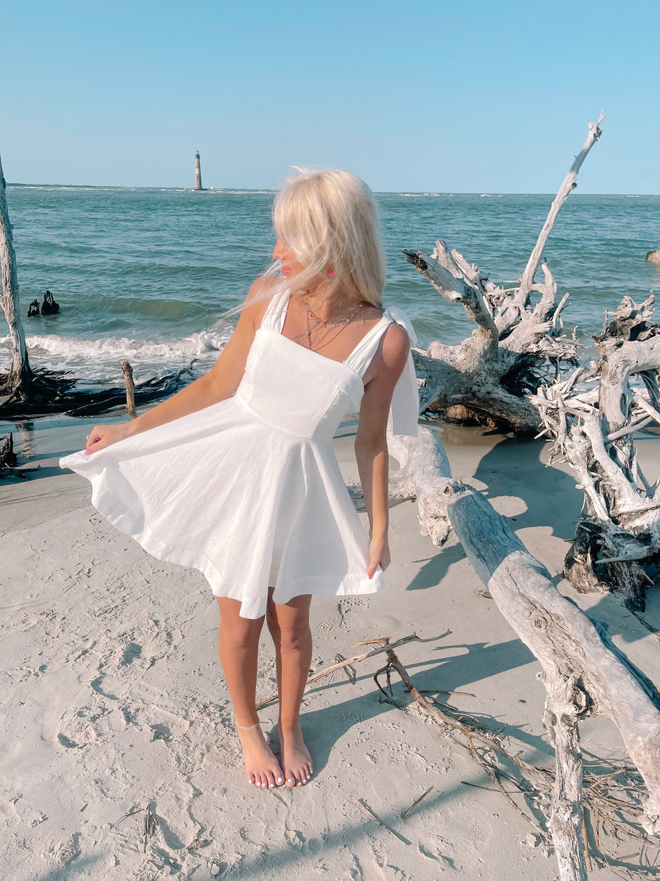 White sundress on beach, near driftwood and ocean.
