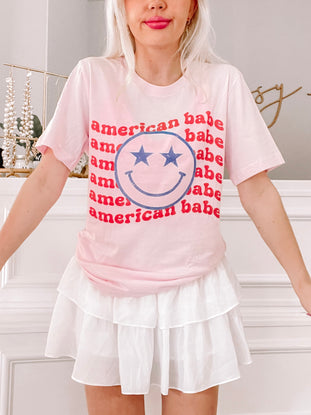 Pink t-shirt featuring a smiley face and repeated "American Babe" text, paired with a white tiered skirt.
