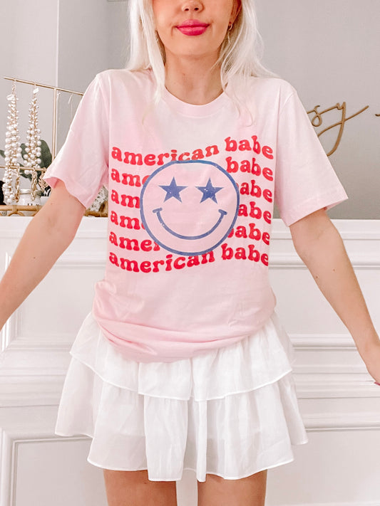 Pink t-shirt featuring a smiley face and repeated "American Babe" text, paired with a white tiered skirt.