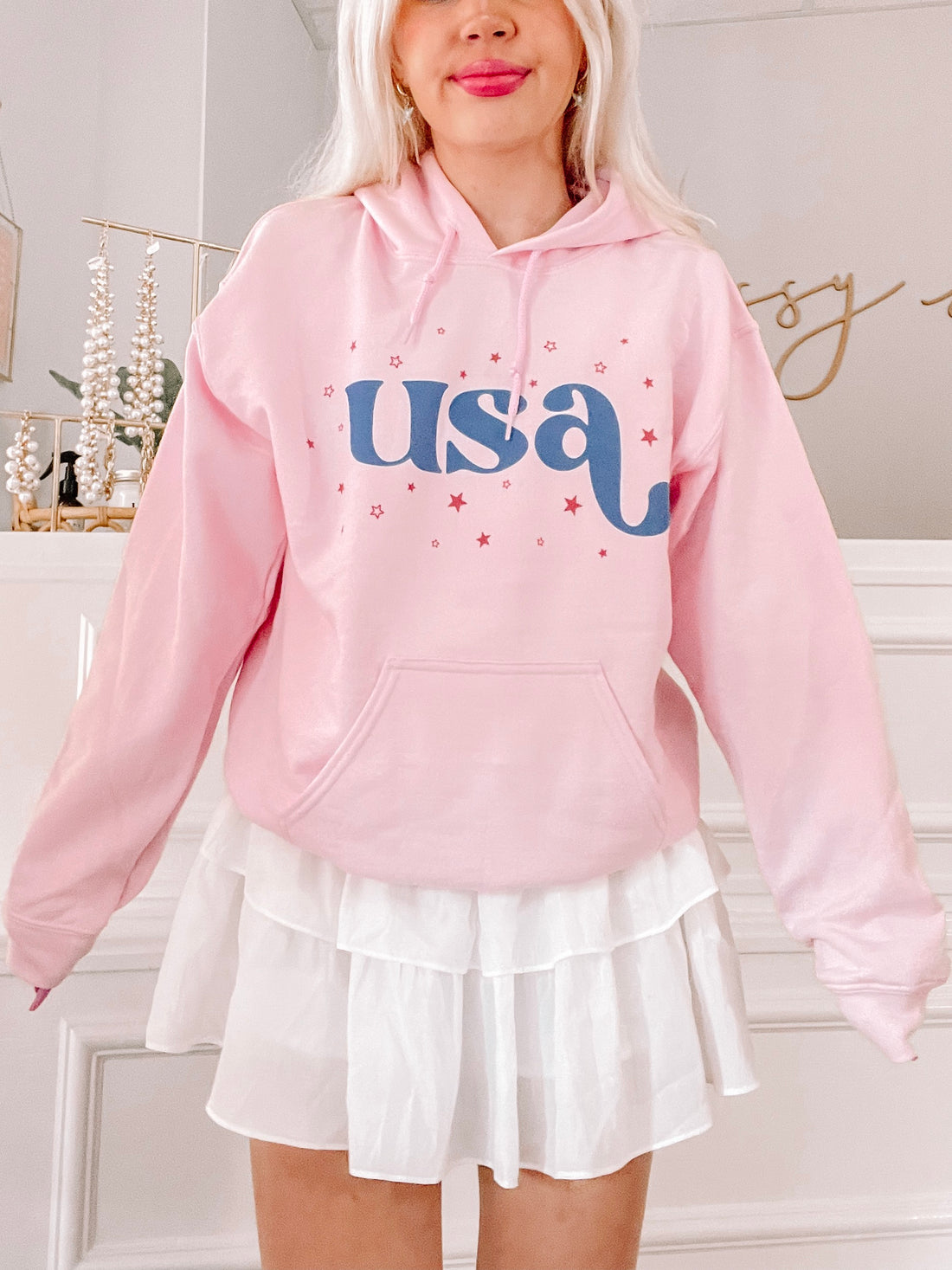 Pink hoodie featuring a retro "USA" graphic and scattered stars, modeled against a light background.
