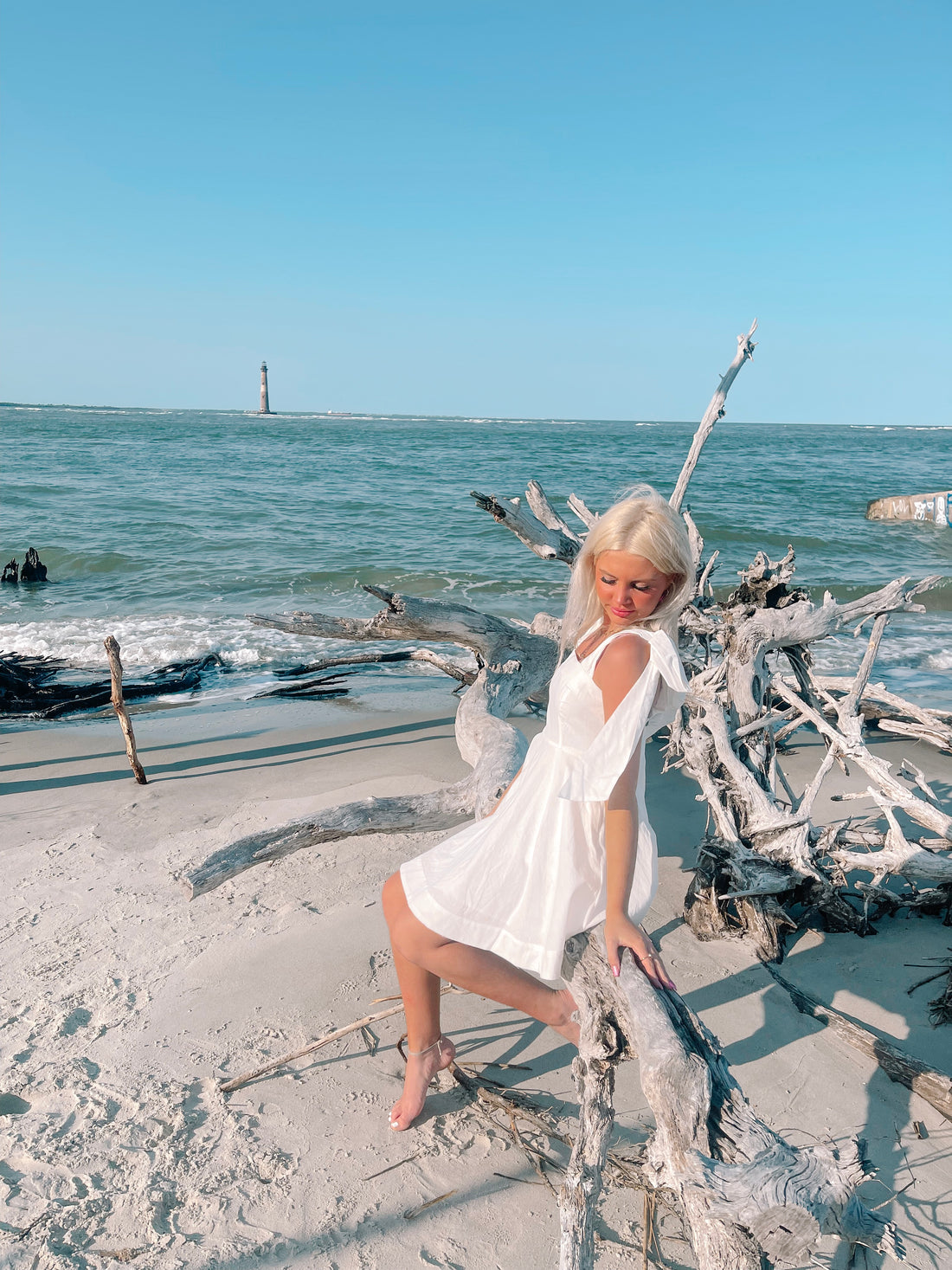 Woman in white sundress sits on driftwood at the beach, ocean and lighthouse in background.
