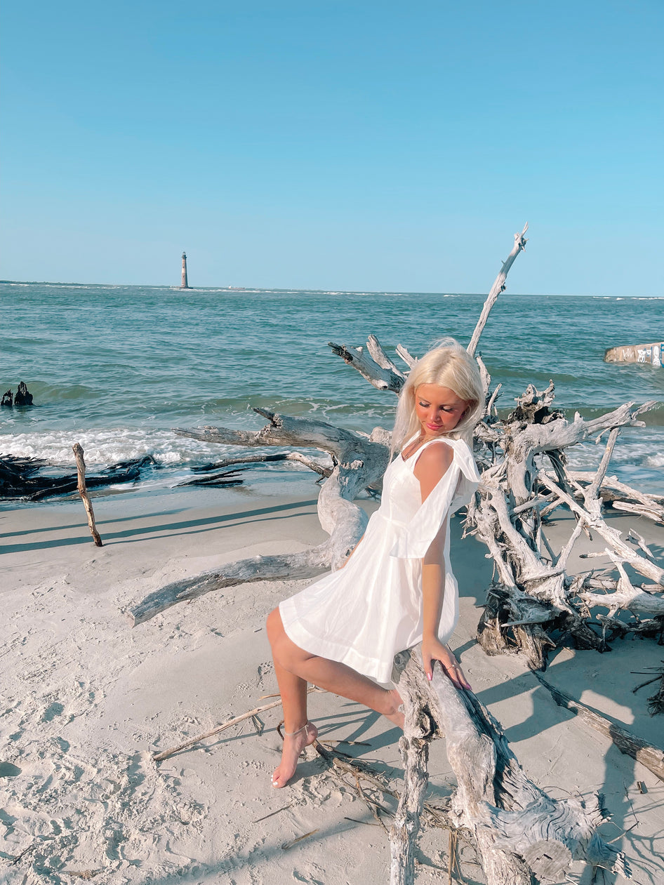 Woman in white sundress sits on driftwood at the beach, ocean and lighthouse in background.
