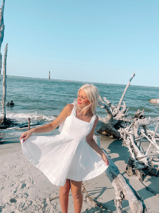 White sundress on beach; girl twirling near driftwood, ocean view.