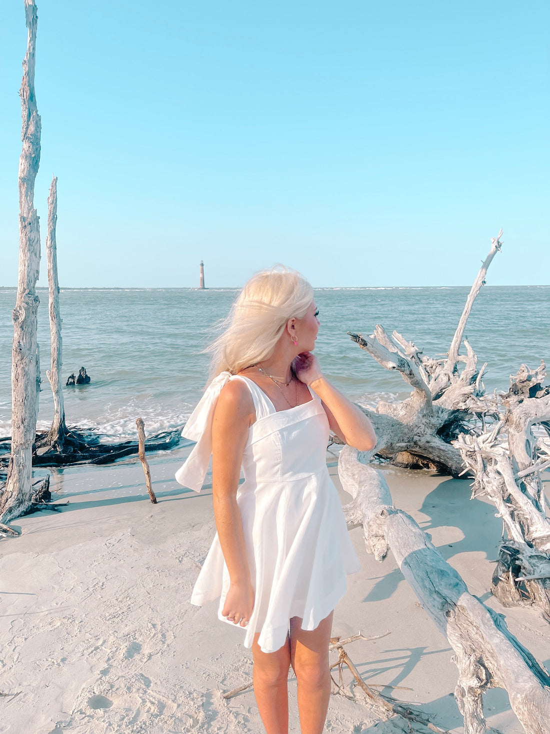 White eyelet dress on a beach, near driftwood and a distant lighthouse.
