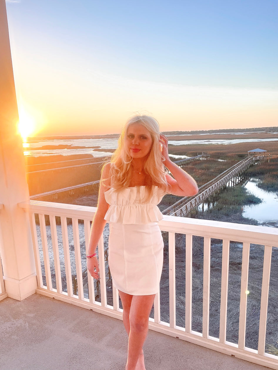 Woman in white mini dress on deck overlooking sunset marsh.
