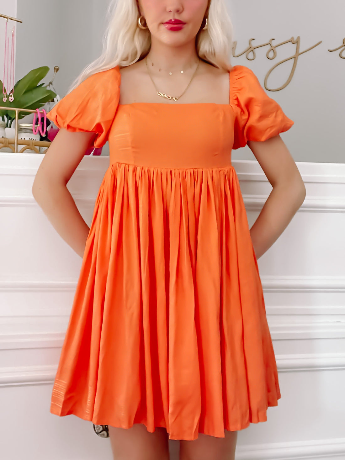 Bright orange mini dress with puff sleeves and square neckline, shown on a model against a white wall.
