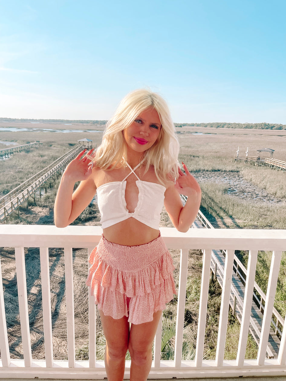 Pink tiered mini skirt with delicate print, modeled outdoors on a porch overlooking a marshy landscape.
