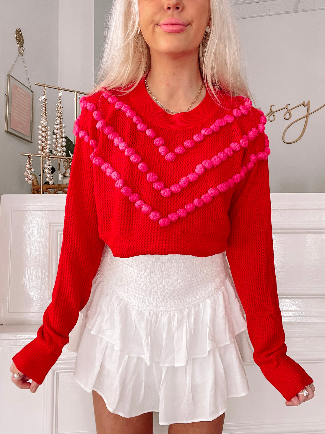 Red waffle-knit sweater with pink pom-pom accents, modeled with white ruffled skirt.
