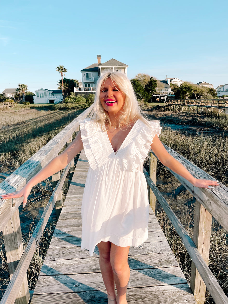 White eyelet mini dress on a wooden boardwalk, sunny coastal background.
