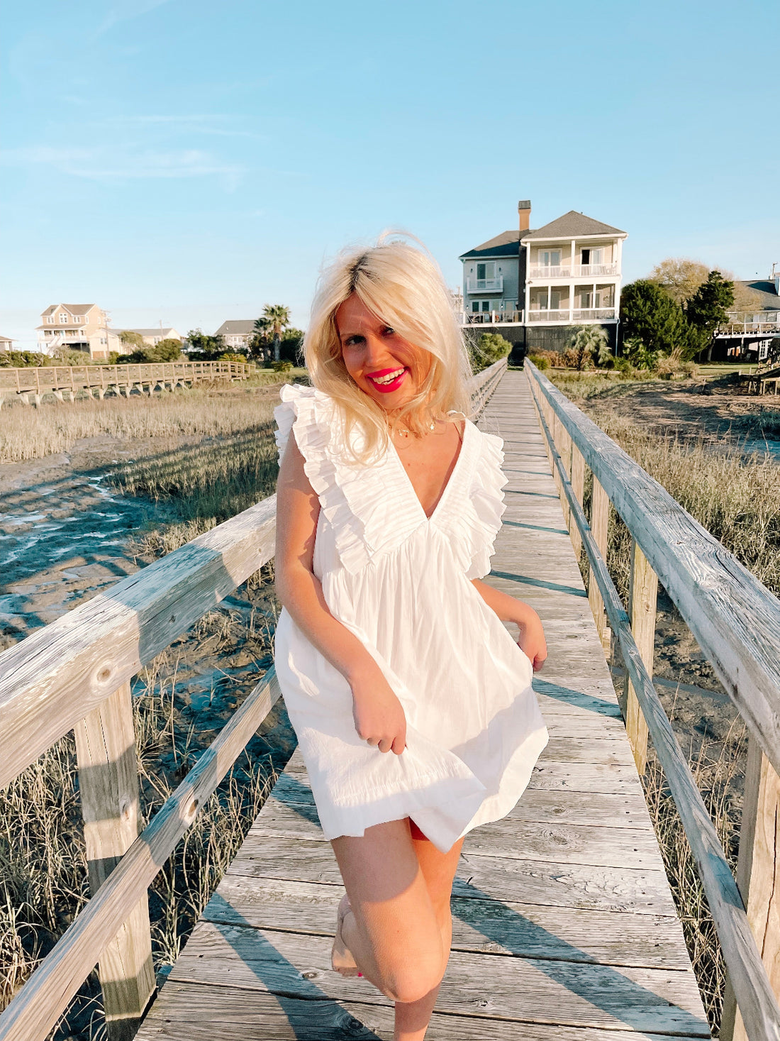 Woman in white ruffled sundress walking on boardwalk near beach homes. 