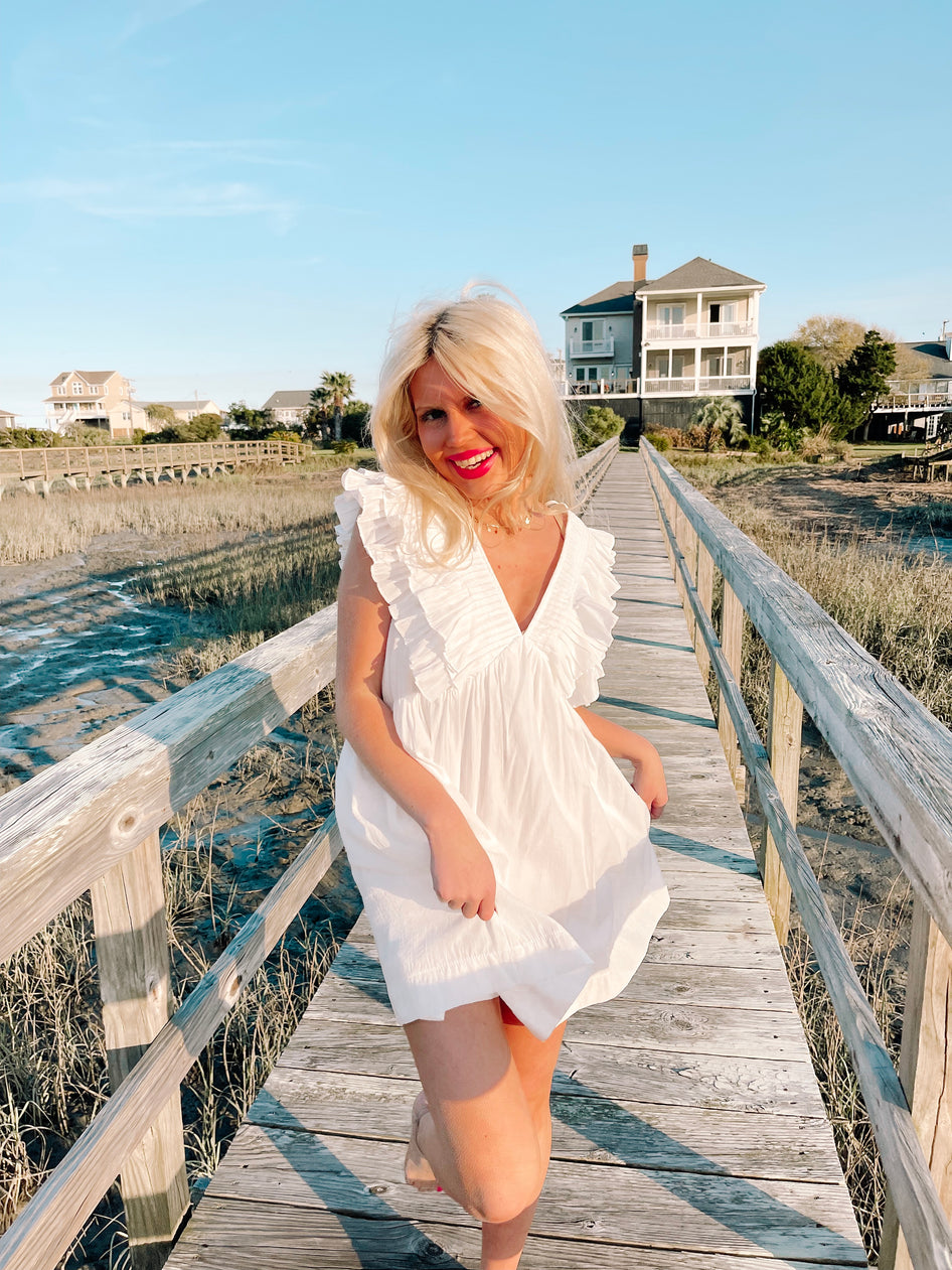 Woman in white ruffled sundress walking on boardwalk near beach homes.
