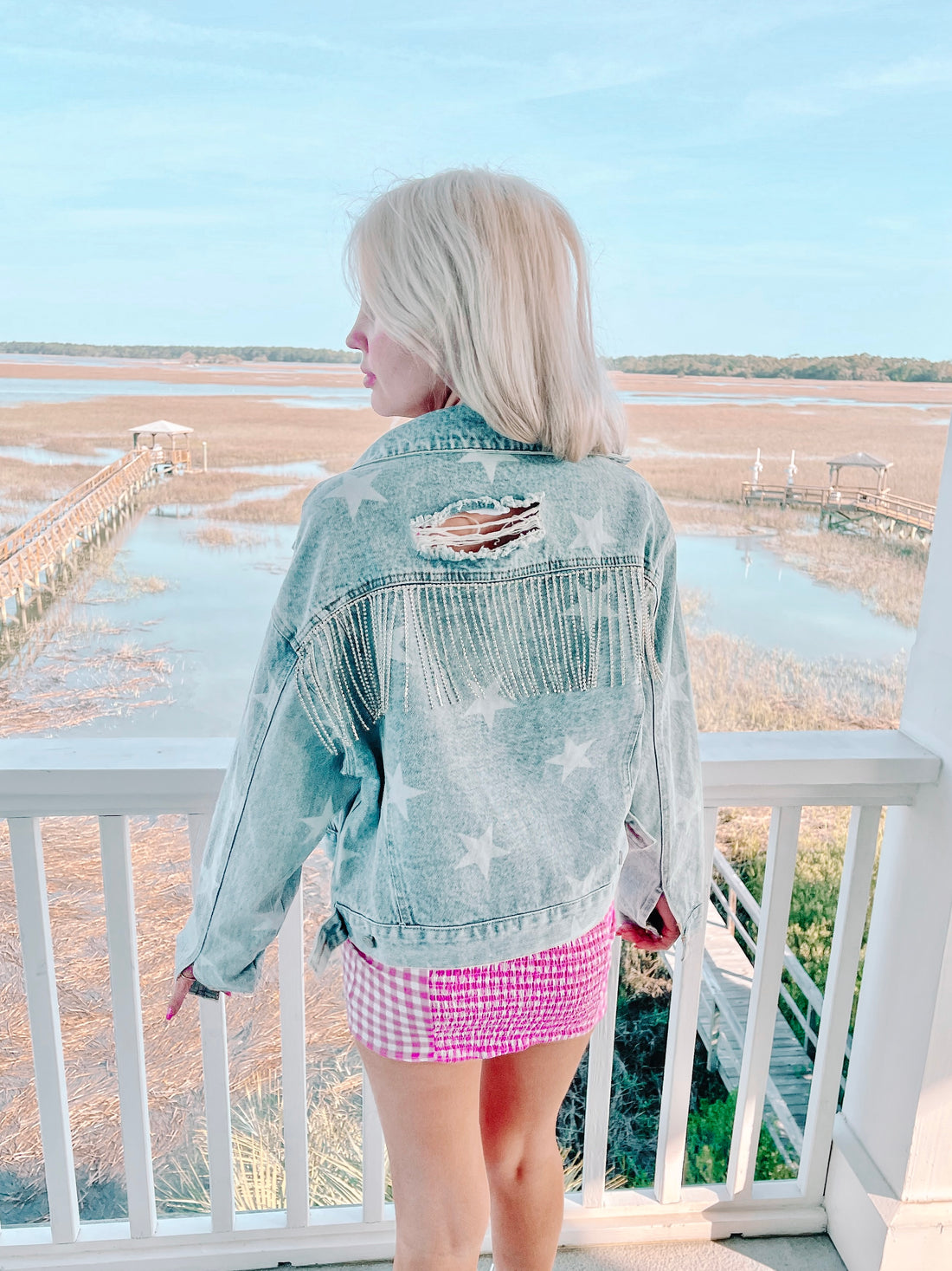 Light wash denim jacket with star details and crystal fringe, viewed from behind on a balcony overlooking a tranquil waterfront.
